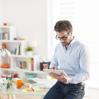 Un homme consulte une tablette tactile, appuyé contre son bureau dans un espace de travail lumineux.