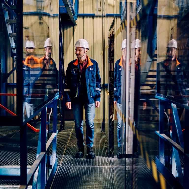 Un technicien en casque et veste de travail marche dans une installation industrielle, entouré de reflets vitrés.