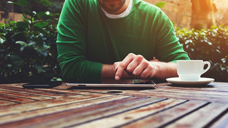 Homme utilisant une tablette tactile sur une table en extérieur, accompagné d'un café